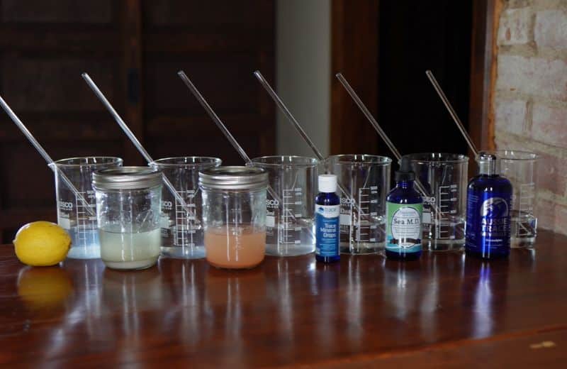 A lineup of clear beakers and jars filled with various colored liquids, each containing a metal stirring rod. In front are three bottles labeled 'Trace Mineral Drops,' 'Sea M.D.,' and 'Aussie Trace Minerals,' alongside a lemon on a wooden table. The scene suggests a DIY experiment or demonstration involving mineral drops and water mixtures