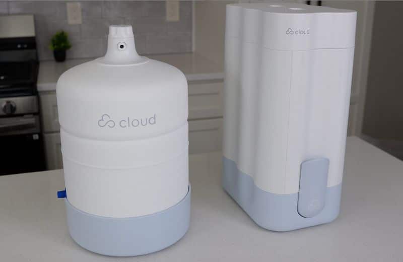 Two white Cloud RO appliances sit on a kitchen countertop. The left unit is the purified water holding tank. The right unit is the reverse osmosis system body. Both display the Cloud RO branding. A stove, backsplash tiles, outlets, and a small plant are visible in the background.