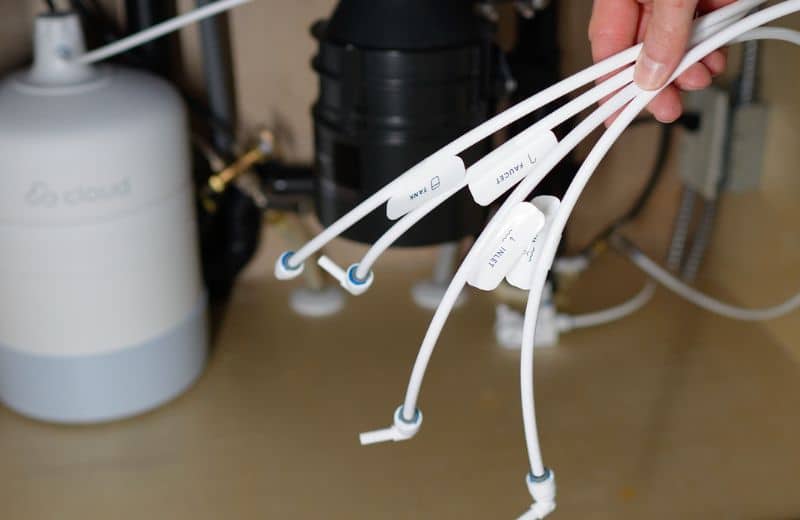 A person holds several labeled white plastic tubes—“FAUCET,” “TANK,” and “INLET”—under a kitchen sink. The tubes are part of a Cloud RO water filtration system. A cylindrical Cloud RO tank, garbage disposal unit, and plumbing connections are visible in the background.