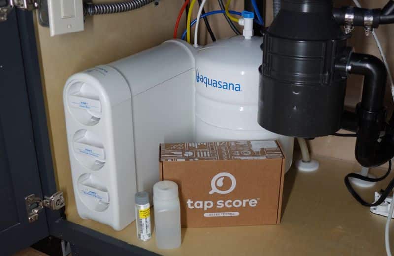 Aquasana SmartFlow Design Undersink Under a kitchen sink, an Aquasana SmartFlow reverse osmosis system with a white tank and three filter cartridges is installed. Beside it is a brown Tap Score water testing box with two sample containers. Pipes, hoses, and wiring surround the setup.