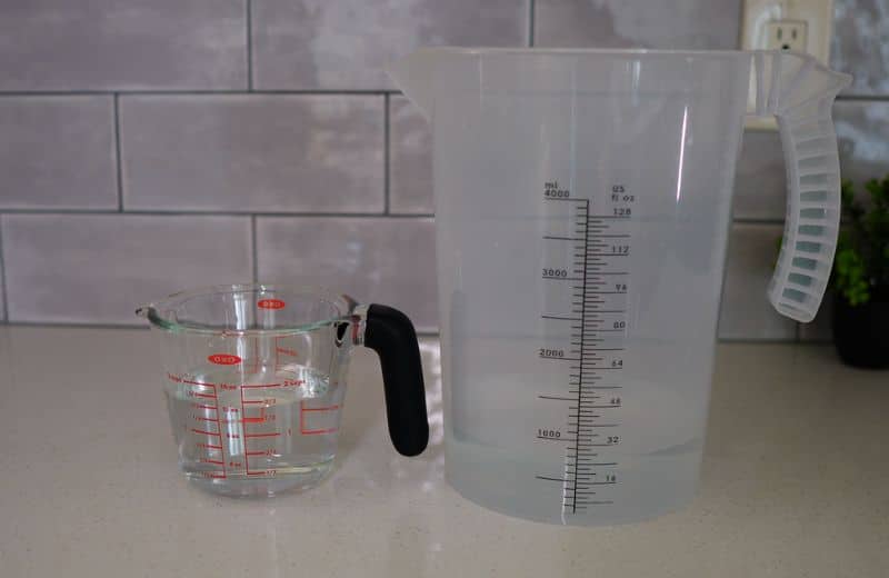 Aquasana SmartFlow Efficiency Ratio On a kitchen counter, a small glass measuring cup with red markings and a black handle sits next to a large plastic jug marked in milliliters and fluid ounces. Showing Aquasana SmartFlow RO efficiency ratio