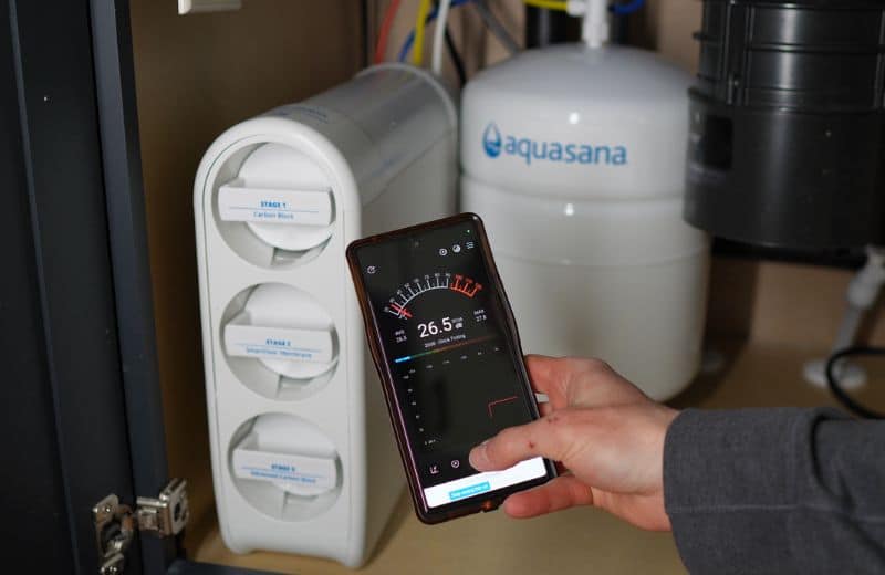 Aquasana SmartFlow Noise Level An Aquasana SmartFlow RO system under a sink shows three labeled filter stages and a white pressurized tank. A person holds a smartphone displaying a conductivity reading of 26.5 dB