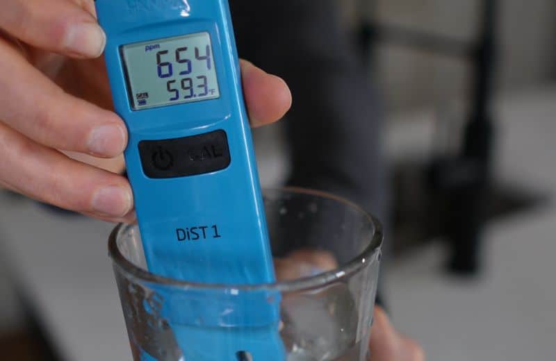 Aquasana SmartFlow TDS A hand holds a blue digital water quality tester in a glass of water to measure Aquasana SmartFlow RO TDS. The display reads 654 ppm and 59.3°F. Buttons labeled “CAL” and “power” are visible.