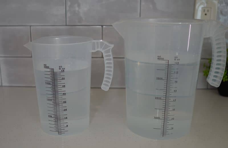 Aquasana Smartflow Efficiency Ratio 1 Two transparent plastic measuring jugs sit on a kitchen countertop. Both have ribbed handles for grip. It shows Aquasana SmartFlow RO efficiency ratio.