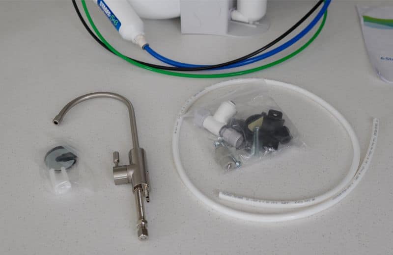 Kind Water PRO Accessories A curved stainless steel faucet sits on a white countertop surrounded by mounting hardware, including rubber and metal washers, a spacer, a lock washer, and a keyhole-shaped plate. Flexible tubing from the KIND WATER PRO RO setup appears behind the parts.