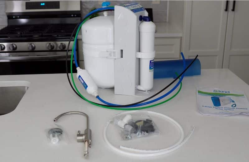 Kind Water PRO Product Kit A 6‑stage KIND WATER PRO RO filtration system sits on a kitchen counter with its white tank, filter cartridges, and blue, green, and black tubing. In front are installation components including a faucet, plastic fittings, and small hardware pieces beside the user manual.