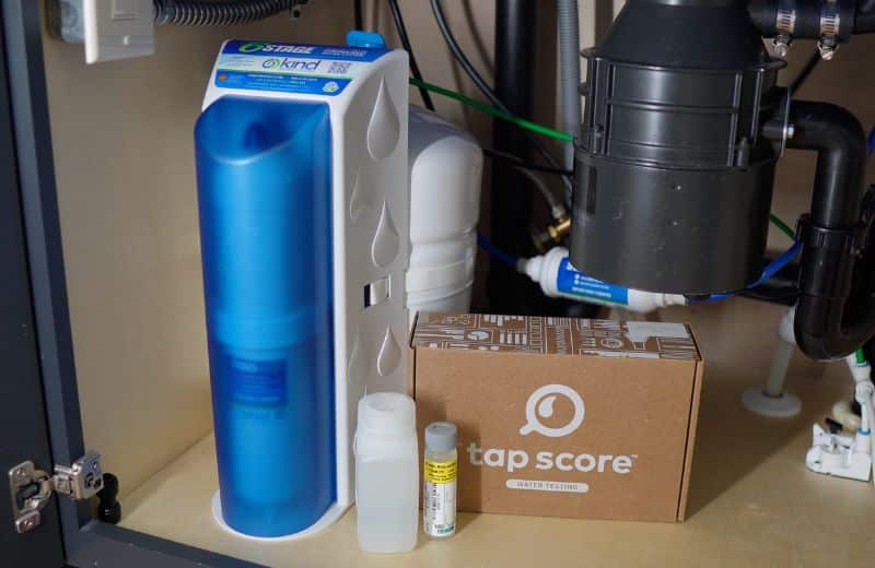 Kind Water PRO Undersink Under a kitchen sink, a KIND WATER PRO RO system with a white tank and blue tubing is installed beside a cardboard box labeled “tap score WATER TESTING.” Two sample containers sit nearby, showing a complete water quality monitoring setup.