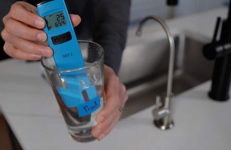 Kind Water second draw TDS Reading A person holds a blue digital water quality tester inside a glass labeled “Prod.” The display shows readings “25” and “654.” A kitchen sink is visible in the background. Part of the KIND WATER PRO RO testing process.