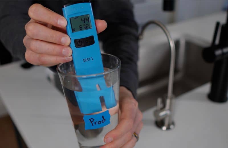KindWater PRO First draw sample TDS A blue DiST 1 digital meter is submerged in a glass labeled “Prod,” displaying “44” and “67.2.” The scene takes place near a kitchen sink as part of KIND WATER PRO RO water quality testing.