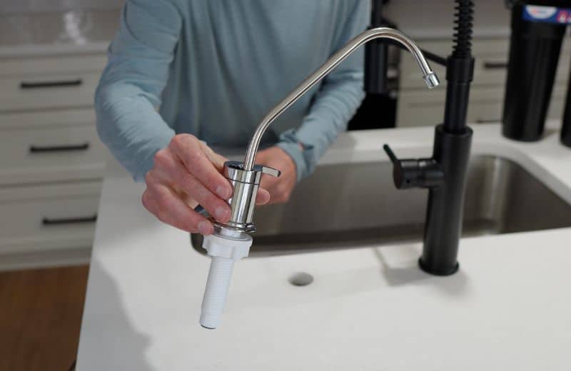 US Water Made RO Faucet Close-up of a polished chrome drinking water faucet from American Water’s RO system being held above a kitchen countertop during installation, next to a matte black sink faucet and under-sink filtration setup.