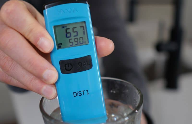 US Water Made RO TDS Initial reading A hand holds a blue digital water quality tester in a glass of water to measure US Water American RO TDS. The display reads 657 ppm and 59°F. Buttons labeled “CAL” and “power” are visible.