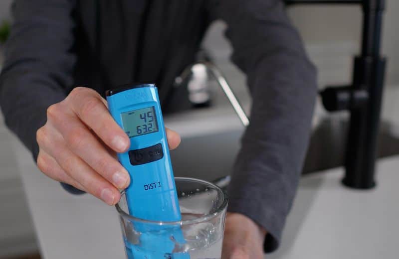 US Water Made RO TDS reading before and after remineralization A hand holds a blue digital water quality tester in a glass of water to measure US Water American RO TDS. The display reads 45 ppm and 63.2°F. Buttons labeled “CAL” and “power” are visible.
