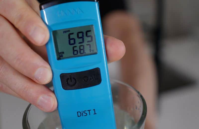 AquaTru Under Sink 2nd Gen Prefiltration TDS A digital water tester reads 695 ppm in a glass of water, illustrating pre‑filtration water quality before passing through the Aquatru Under Sink RO 2nd gen system.