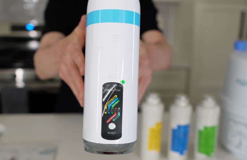 AquaTru Under Sink 2nd Gen Unit Buttons A cylindrical water filter unit with a colored indicator panel is held upright, shown alongside replacement cartridges compatible with the Aquatru Under Sink RO 2nd gen system.