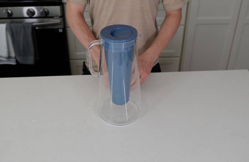 LifeStraw Home Pitcher Design A person holds a clear plastic pitcher with a blue lid and inner cylinder insert at a kitchen counter, demonstrating the design and usability of the LifeStraw Home Essential 10‑Cup Pitcher Review.