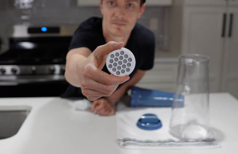 LifeStraw Home Pitcher Filter Material A person in a kitchen holds a circular filter or aerator with multiple holes, shown alongside a pitcher and lid on the counter, highlighting parts of the LifeStraw Home Essential 10‑Cup Pitcher Review.
