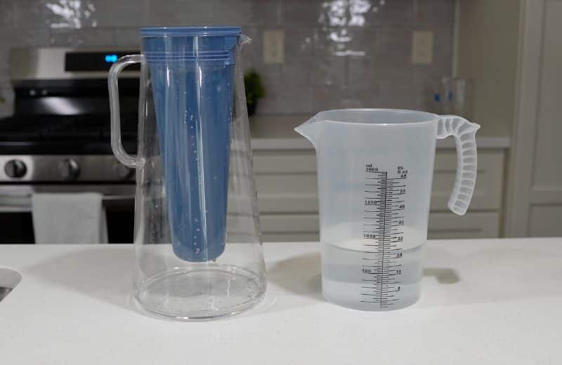 LifeStraw Home Pitcher Water Volume Usable A clear plastic pitcher with a blue cylindrical insert sits beside a translucent measuring jug filled with water on a kitchen counter, illustrating water filtering and measuring in the LifeStraw Home Essential 10‑Cup Pitcher Review.